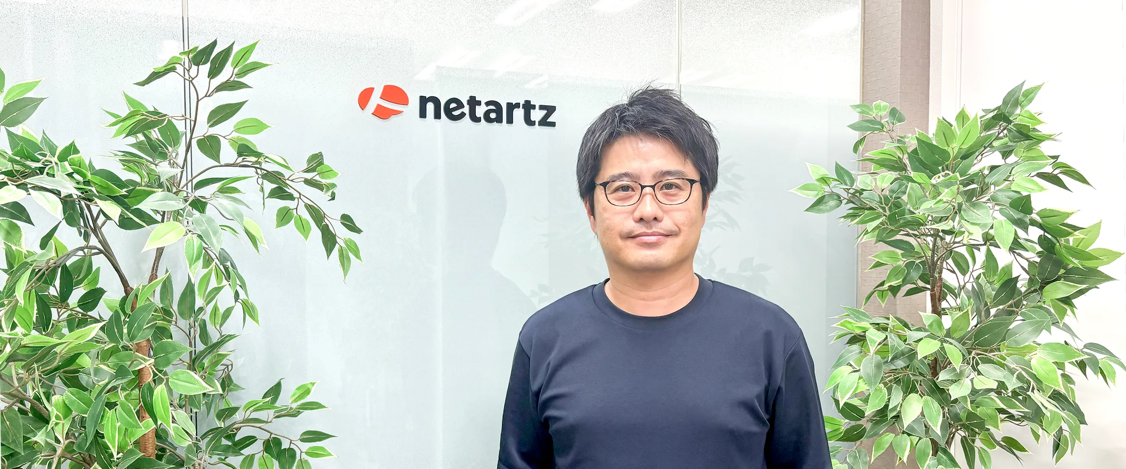 株式会社ネットアーツのオフィスでロゴの前に立つ生井文夫様