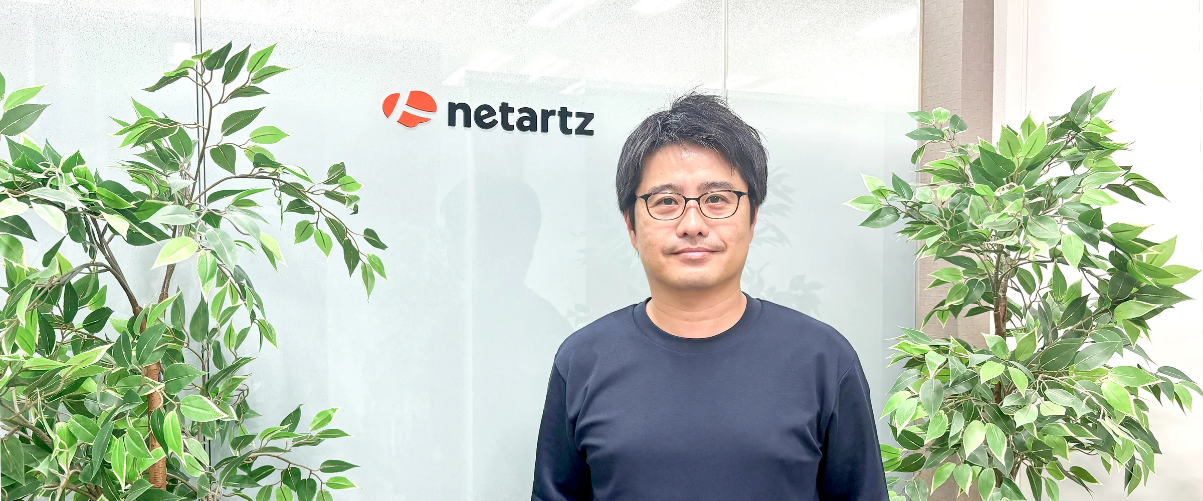 株式会社ネットアーツのオフィスでロゴの前に立つ生井文夫様